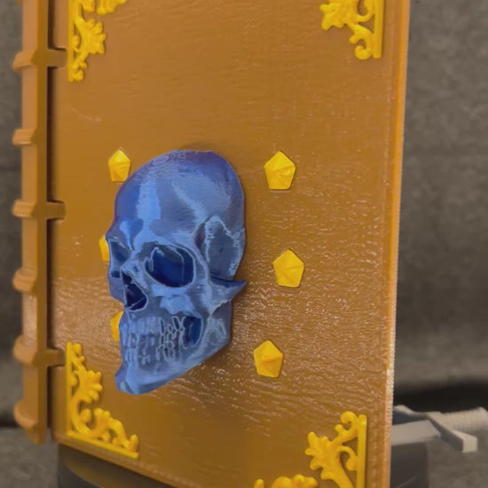 The Sworn Grimoire deck box turning on a display stand, showing the skull-and-sword cover and gold tooling.
