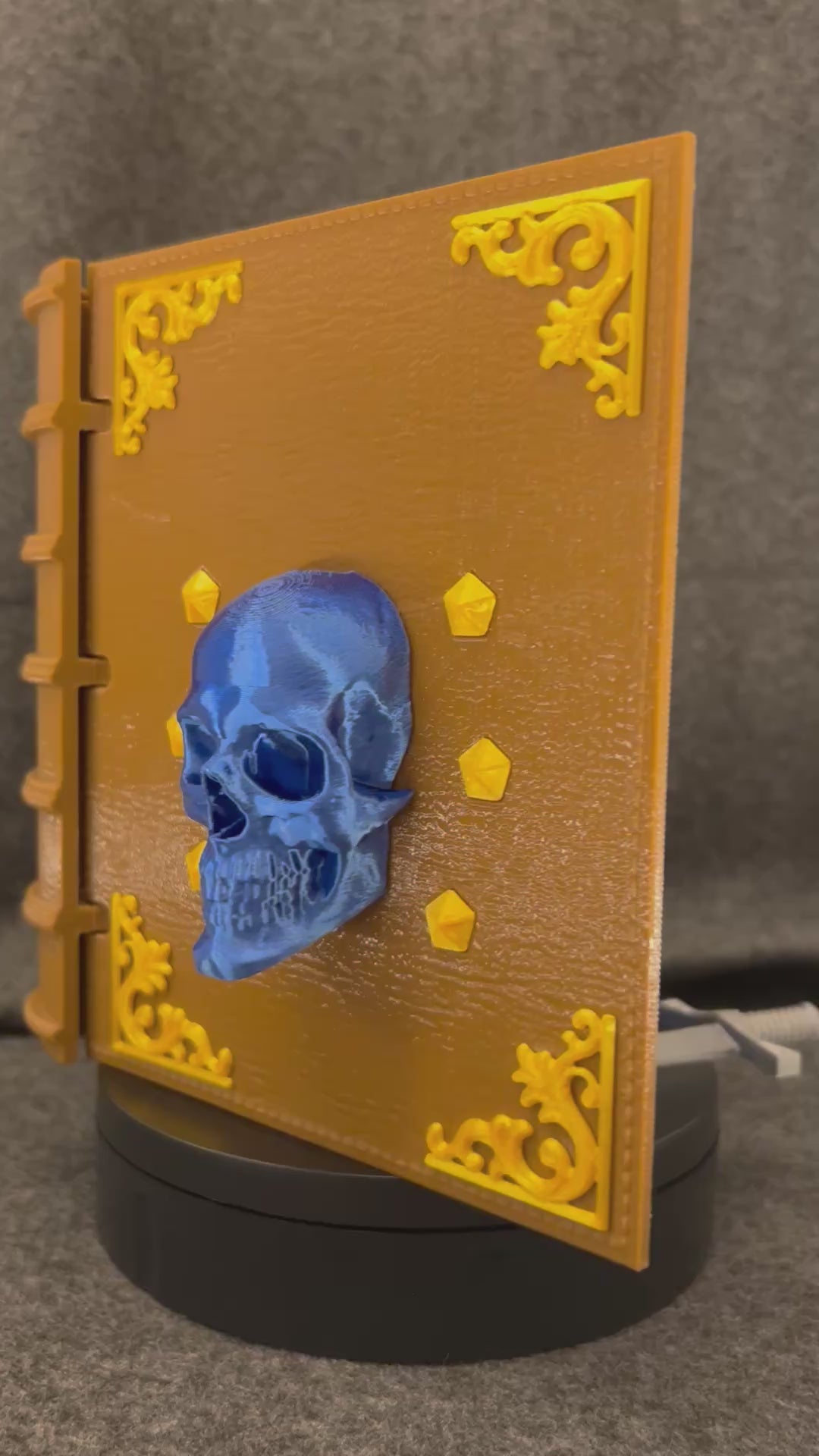 The Sworn Grimoire deck box turning on a display stand, showing the skull-and-sword cover and gold tooling.